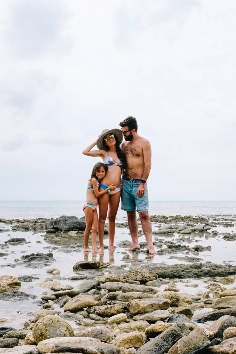 Drea, husband, and daughter standing together in front of beach